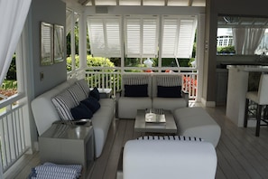 A covered outdoor seating area with a white sofa, striped cushions, and a small table. - Tradewinds at Sandy Lane Estate | Timeless Luxury in Barbados’ Most Prestigious Address (Holetown)