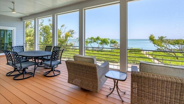 A covered outdoor area with a table and chairs, large windows offering a view of the sea and greenery.