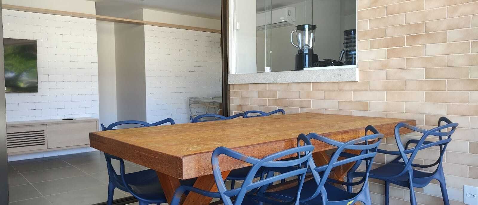 Een eetruimte met een houten tafel en blauwe stoelen, een witte bakstenen muur en een keuken met een blender.