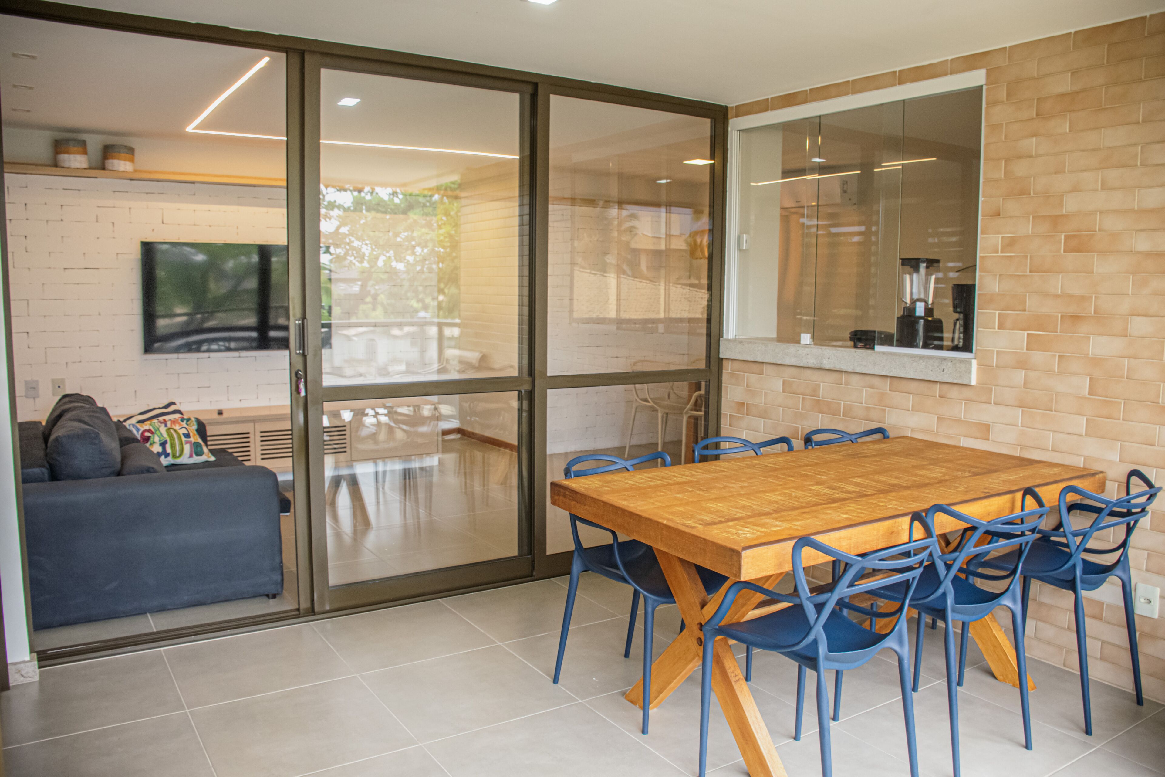 A modern dining area with a wooden table and blue chairs, a sofa, and a TV mounted on a brick wall.