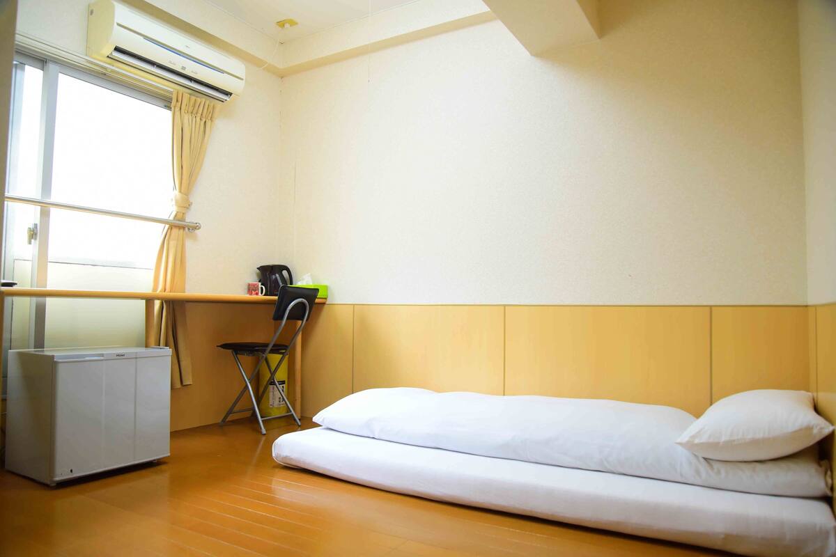 A small, clean room with a bed, a desk, and an air conditioning unit.