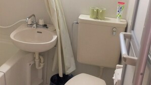 Un baño pequeño y funcional con lavamanos, inodoro y ducha blancos. Hay dos tazas verdes sobre el tanque del inodoro y un cubo de basura junto al inodoro.