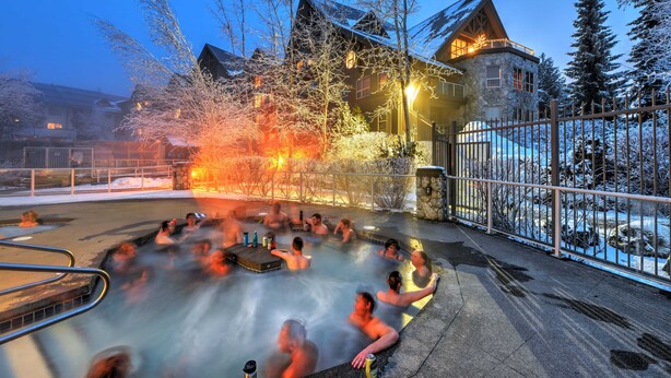A hot tub area with people relaxing, surrounded by snow-covered trees and a stone building with a wooden roof. - Aspens #439 Prime Ski-in Ski-out Location! Top Floor Unit, Pool, Hot tubs, BBQ (Whistler)
