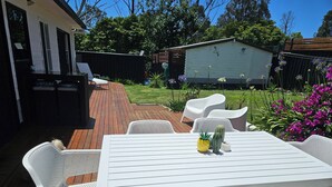An outdoor patio with a white table and chairs, surrounded by a wooden deck and a garden with various plants and flowers.
