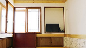 Ein Zimmer mit einem Holzschrank, einem Fernseher auf einem Standfuß und einem Fenster mit Jalousien.