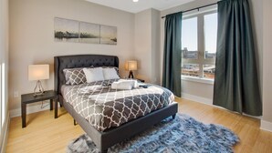 Ein Schlafzimmer mit einem Bett, zwei Nachttischen, einem Fenster mit Vorhängen und Blick auf die Stadt.