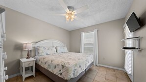 A bedroom with a bed, a nightstand, a ceiling fan, a window with blinds, and a mounted TV. - Sundial G3- The Beach is Where You Need to Be! (Gulf Shores)