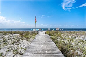 Ein Holzsteg führt zu einem Strand mit einer Flagge, Sonnenschirmen und einem Schild.