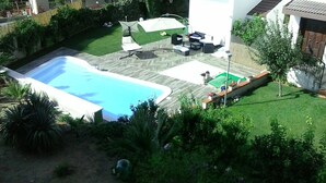 Uma piscina retangular com padrão em forma de cunha ao redor, cercada por uma área gramada com algumas plantas. Há móveis de exterior e um guarda-sol branco ao fundo.