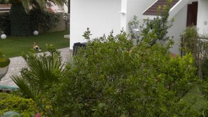 Um edifício branco com persianas castanhas, uma varanda e um jardim com flores cor-de-rosa.
