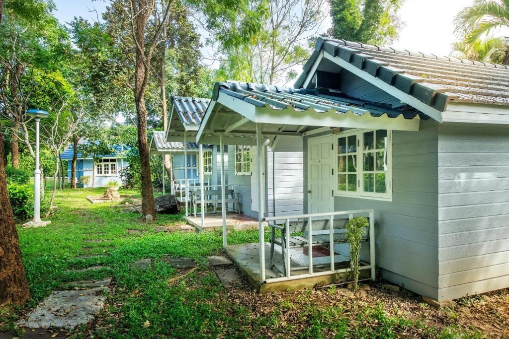 A row of small, light-colored cabins with a covered porch and a tiled roof, surrounded by greenery and trees.