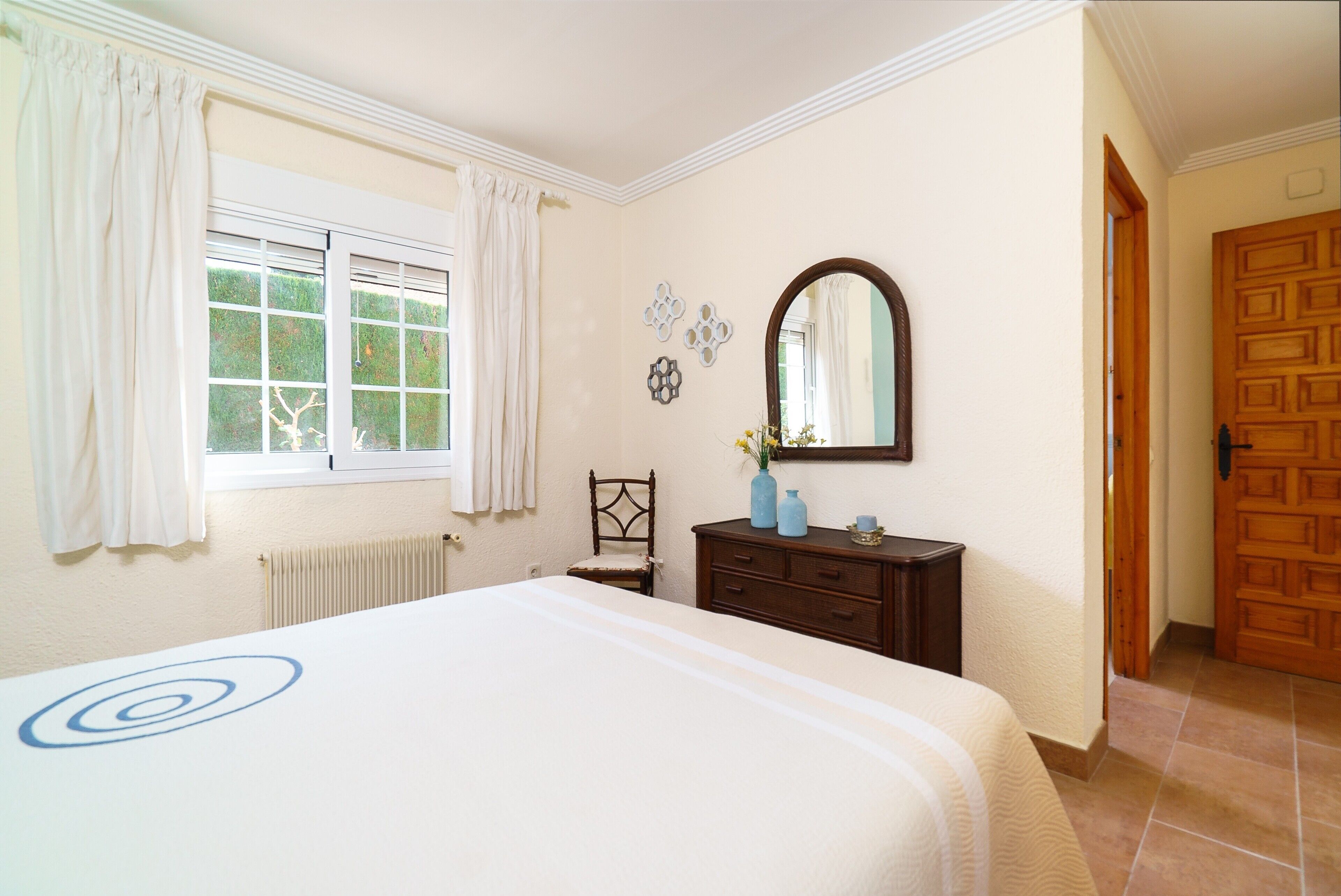 A bedroom with a large bed, a dresser, a chair, a window with curtains, a mirror, and a door.