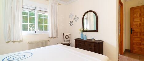 A bedroom with a large bed, a dresser, a chair, a window with curtains, a mirror, and a door.