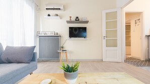 Ein modernes Wohnzimmer mit grauem Sofa, einem Holztisch mit Pflanze, einem Heizkörper, einer Klimaanlage und einem Fernseher auf einem Regal.