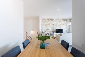 A modern dining area with a wooden table, chairs, a vase with greenery, a small round table, and a TV in the background. - Serene Horizon - BY SCH (Nazaré)