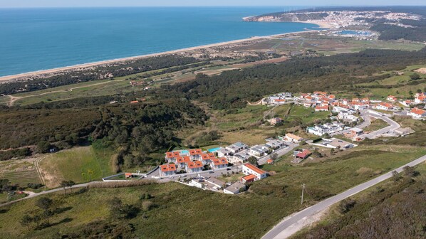 An aerial view of a coastal town with a beach, green fields, and a cluster of buildings with red roofs.