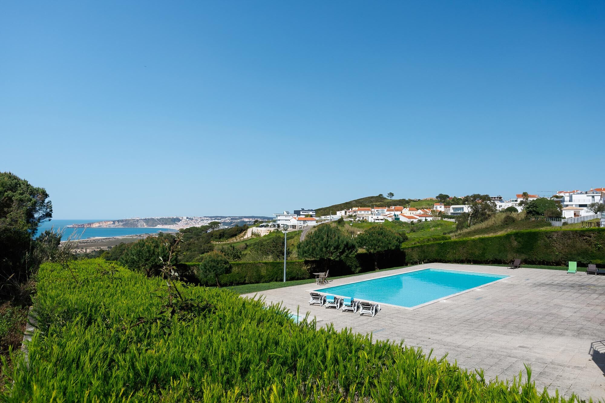 Une piscine entourée de verdure, avec des chaises longues, d’où l’on peut admirer une ville côtière et la mer à l’horizon.