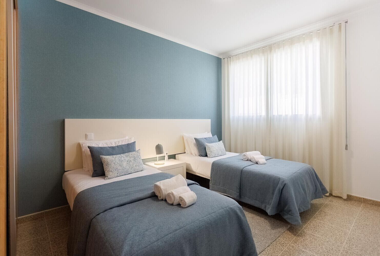 A hotel room with two beds, a nightstand, a lamp, and a window with curtains.