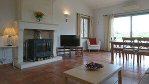 Un salon avec une cheminée, un téléviseur, un coin repas avec une table et des chaises, et une grande fenêtre avec des rideaux.