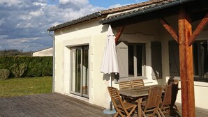 Une terrasse couverte avec une table et des chaises en bois, un parasol blanc et une vue sur une maison avec un toit en tuiles et de grandes fenêtres.