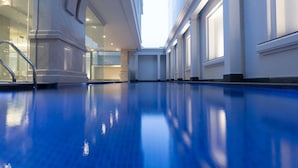 Uma grande piscina interior com piso de azulejos azuis, cercada por paredes brancas e amplas janelas.