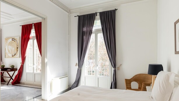 A hotel room with a large bed, a chair, a table with a lamp, and a view of the outside. - Large two bedroom apartment (Barcelona)