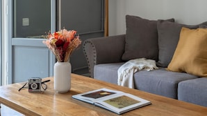 Ein Holztisch mit einer Kamera und einem Buch, ein graues Sofa mit Kissen sowie eine Vase mit getrockneten Blumen.