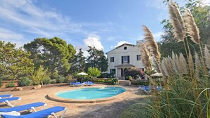 A swimming pool with lounge chairs, surrounded by greenery and a large building in the background.