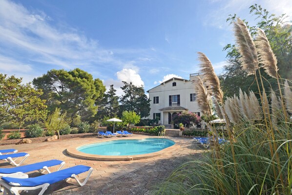 A swimming pool with lounge chairs, surrounded by greenery and a large building in the background.
