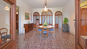 A dining area with a round table, chairs, and a chandelier. There are wooden doors and a potted plant.