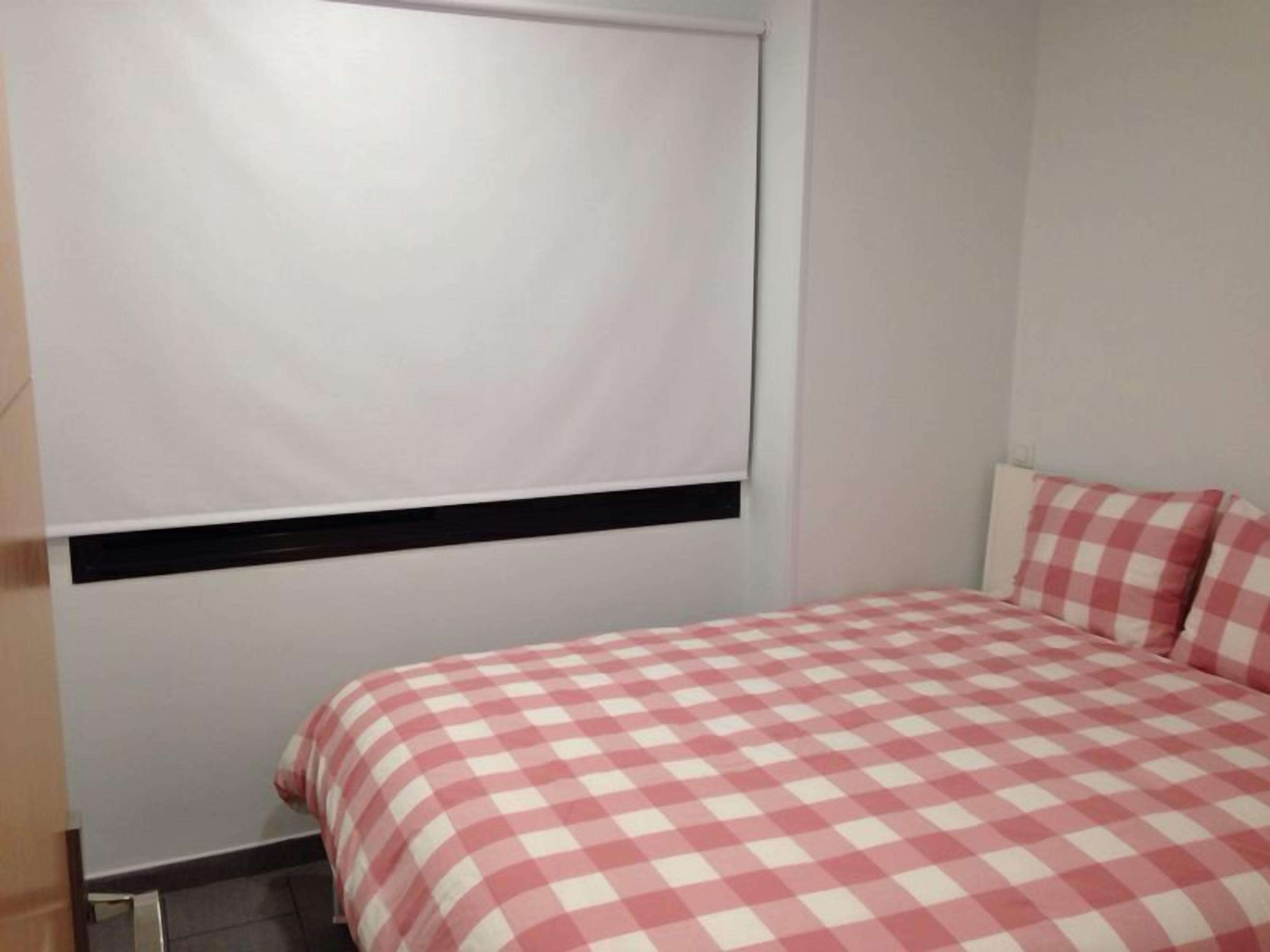 A bedroom with a checkered bedspread, a window with a blind, and a wall-mounted heater.