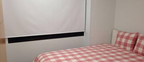 A bedroom with a checkered bedspread, a window with a blind, and a wall-mounted heater.