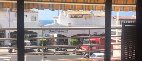 A view from a window with striped blinds, overlooking a street with parked cars and buildings, including a pharmacy and a restaurant.