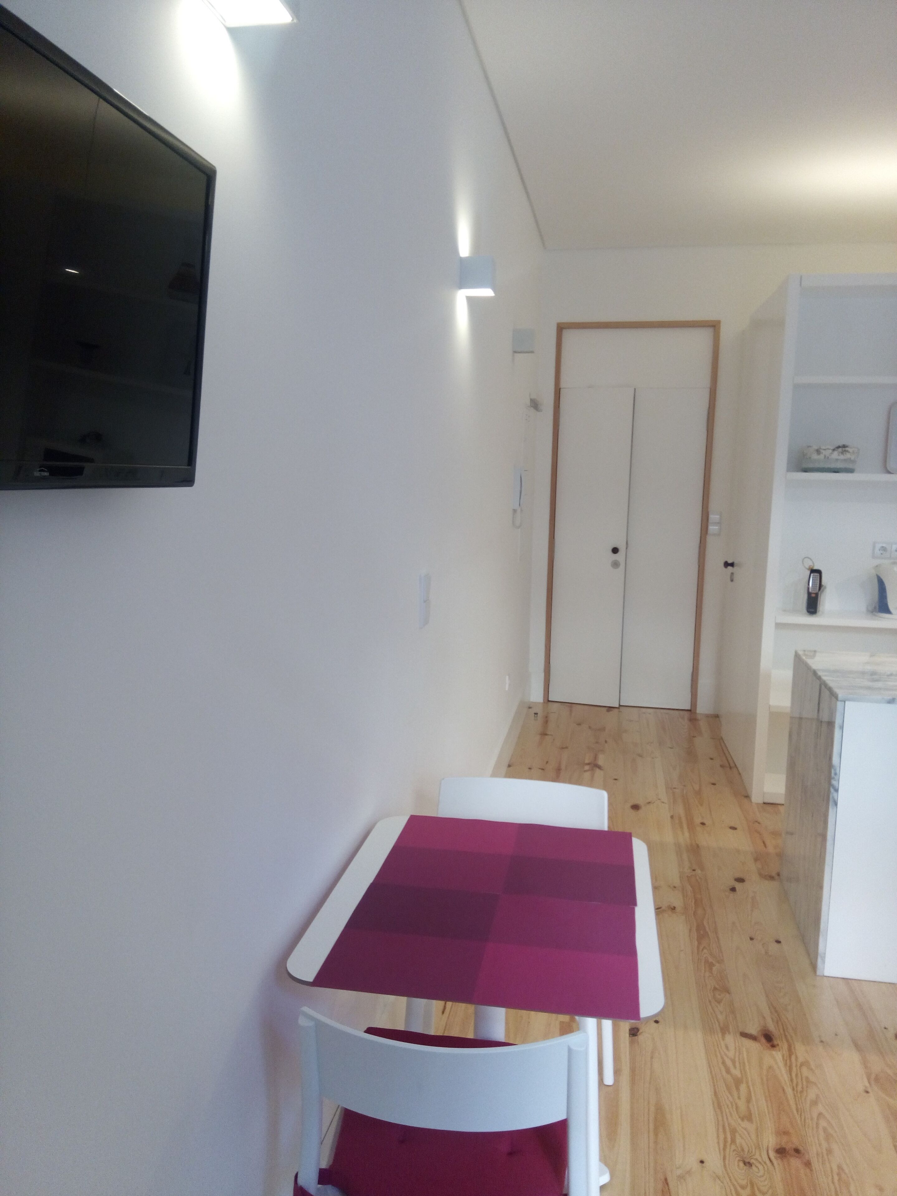 A small room with a white table and chair, a television mounted on the wall, and a door leading to another room.