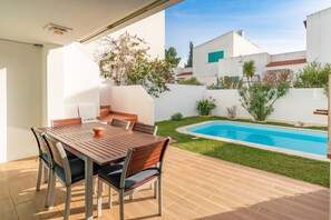 A patio with a wooden table and chairs, a swimming pool, and a white building with a red roof in the background.