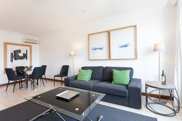 A modern living room with a grey sofa, black chairs, a glass coffee table, and two framed artworks on the wall.