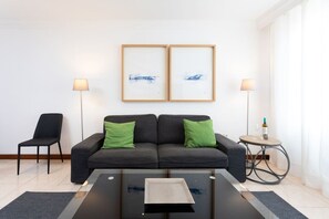 Una sala moderna con un sofá, dos cojines verdes, una mesita de centro, una mesita auxiliar con una botella y vasos, y dos obras de arte enmarcadas en la pared.