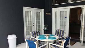 Une terrasse avec une table ronde et six chaises, des portes blanches et une poubelle.