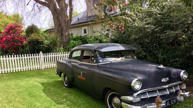 A vintage black car parked on a grassy lawn with a white picket fence and lush greenery in the background. - Rucksacker Backpacker Hostel (Christchurch)