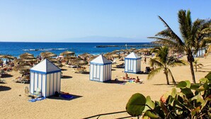 A beach with thatched umbrellas, sun loungers, and small blue and white beach huts.