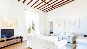 A bright, white-walled room with a wooden ceiling, a television on a wooden stand, a white sofa, and a white armchair.