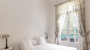 A bedroom with a large bed, white bedding, a nightstand with a lamp, and a window with floral curtains. - Deluxe two bedrooms apartment (Barcelona)
