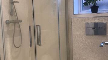 Une douche en verre avec un pommeau de douche à main et un flexible de douche, des toilettes blanches et une fenêtre agrémentée d’une plante en pot.
