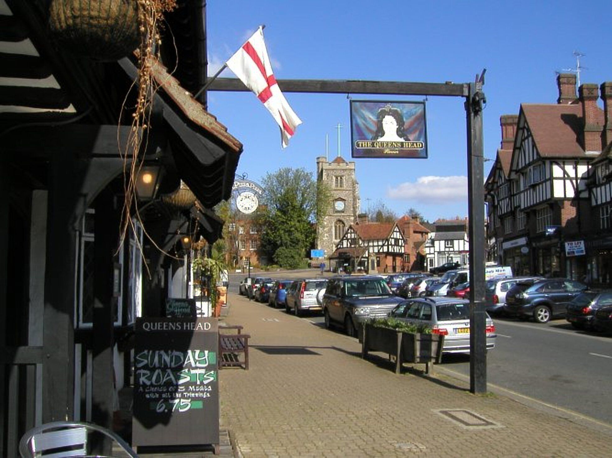 Vista de calle con un pub llamado 'Queens Head' que ofrece asados dominicales.