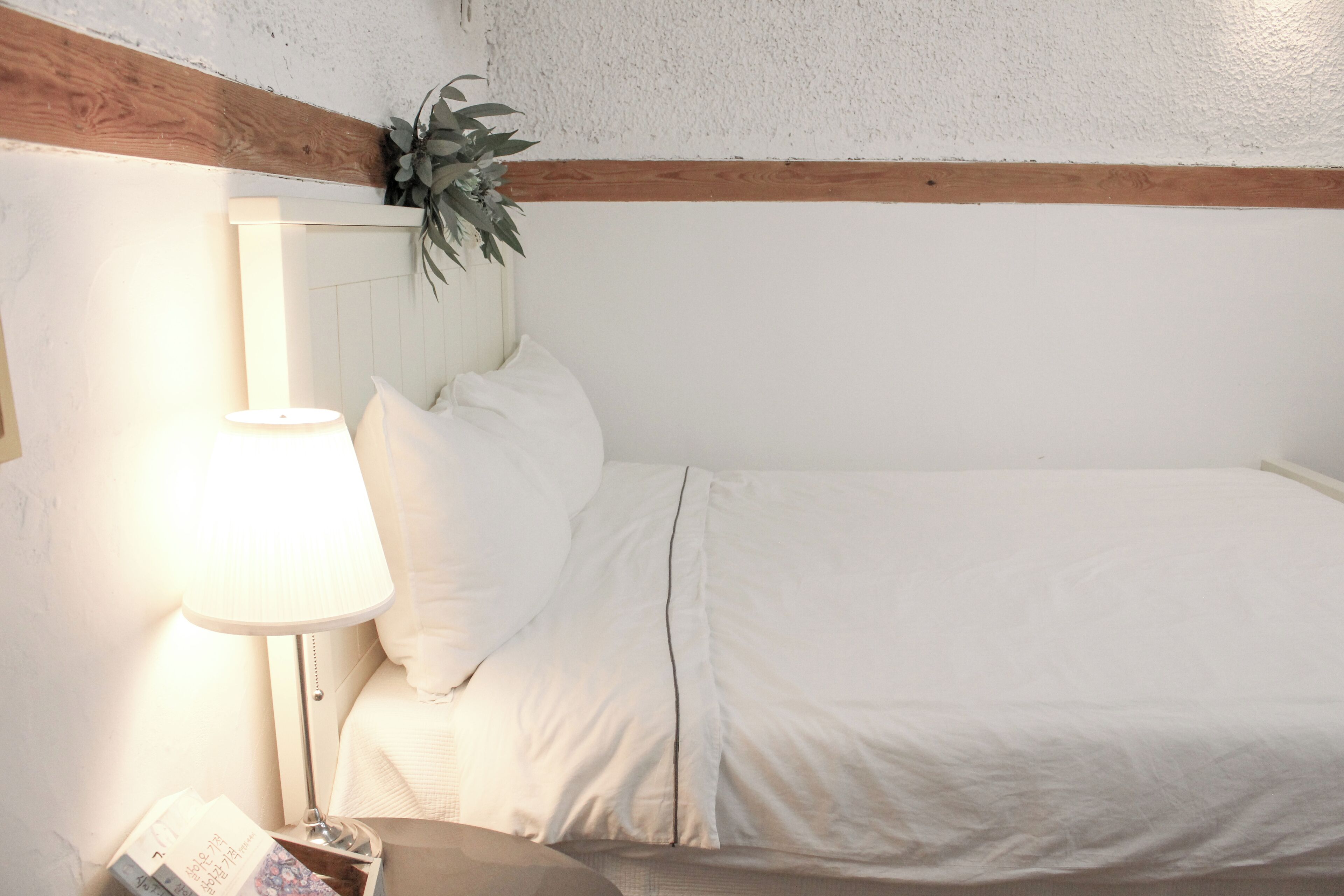 Una cama bien hecha con sábanas blancas, una lámpara de noche y una planta pequeña en la cabecera.