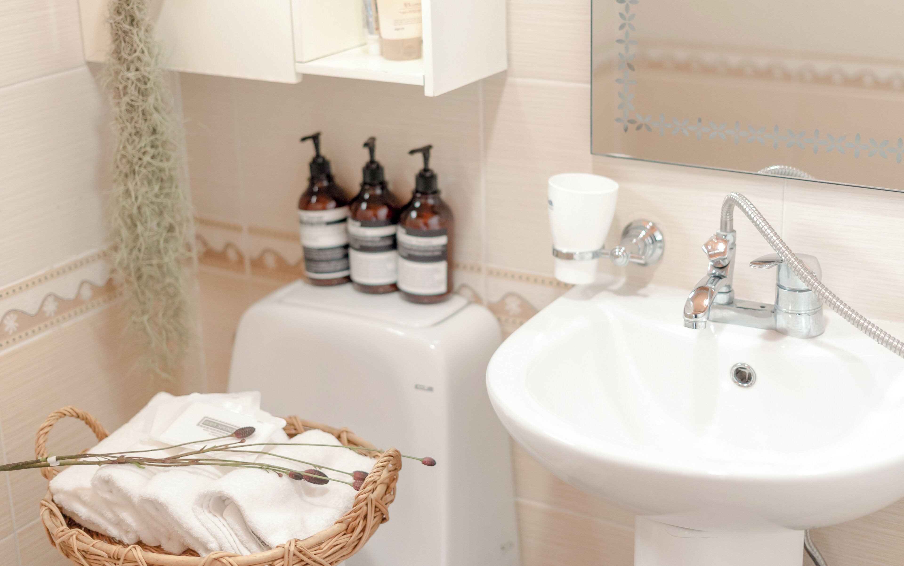 Une salle de bain avec un lavabo blanc, un miroir et un panier rempli de serviettes. Trois bouteilles de savon liquide sont posées sur une étagère au-dessus du lavabo.