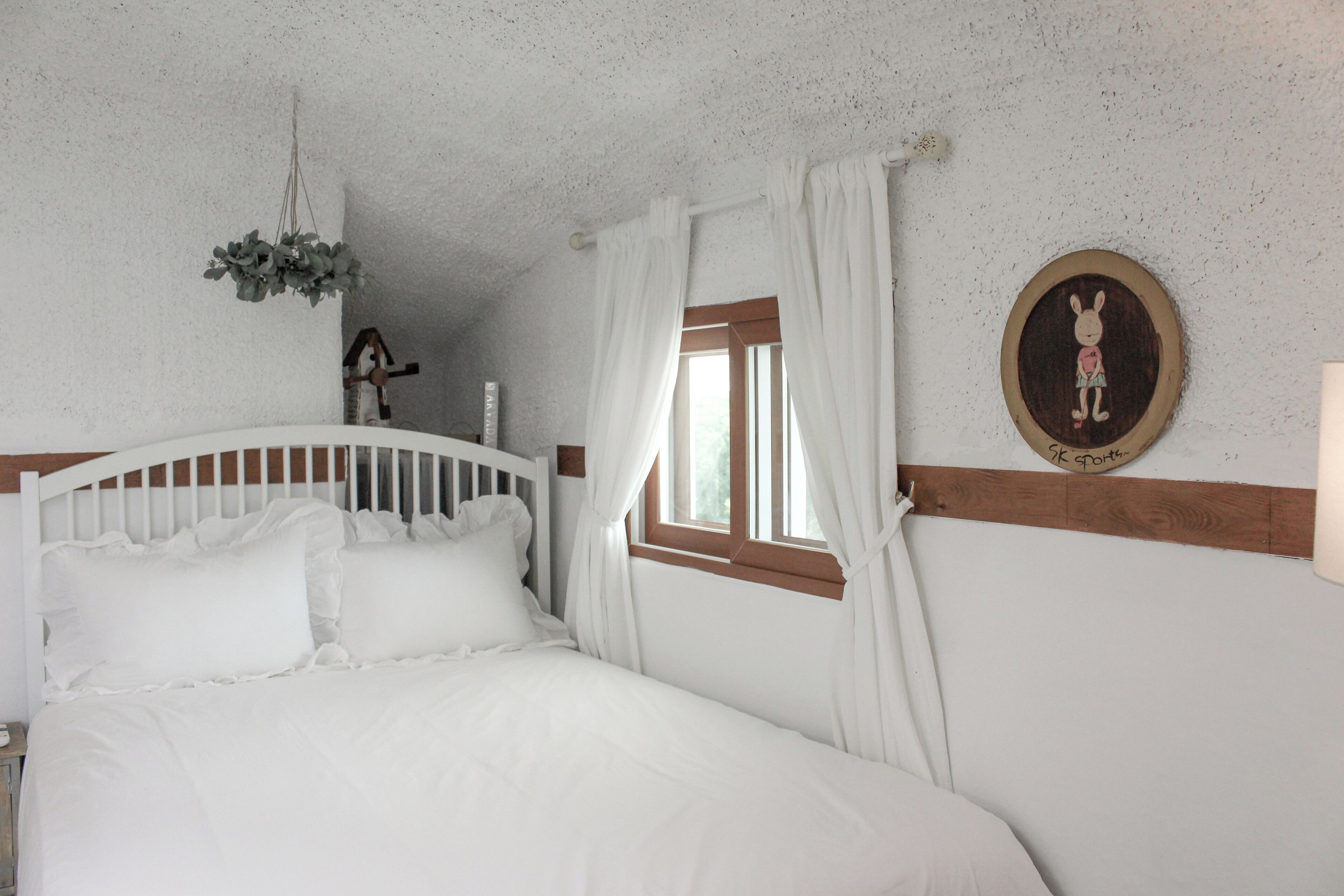 Una cama blanca con cabecera de madera, cortinas blancas y una ventana pequeña con cortinas blancas.