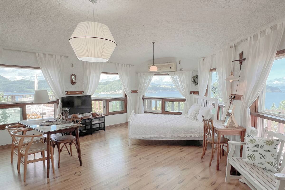 Una habitación con cama, televisión, mesa de comedor y sillas. Hay ventanas con cortinas y vistas al exterior.