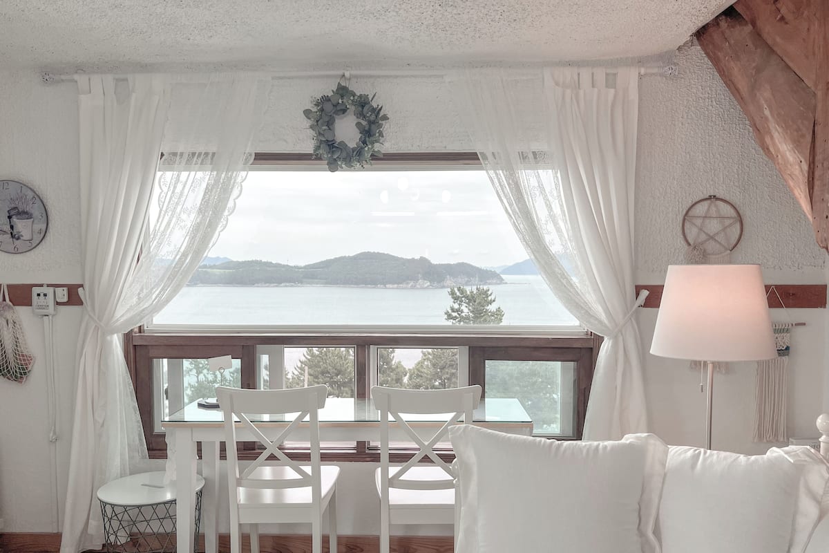 Habitación con vistas a un cuerpo de agua y colinas, que incluye una mesa con sillas, una lámpara y una pared blanca con un reloj y un atrapasueños.