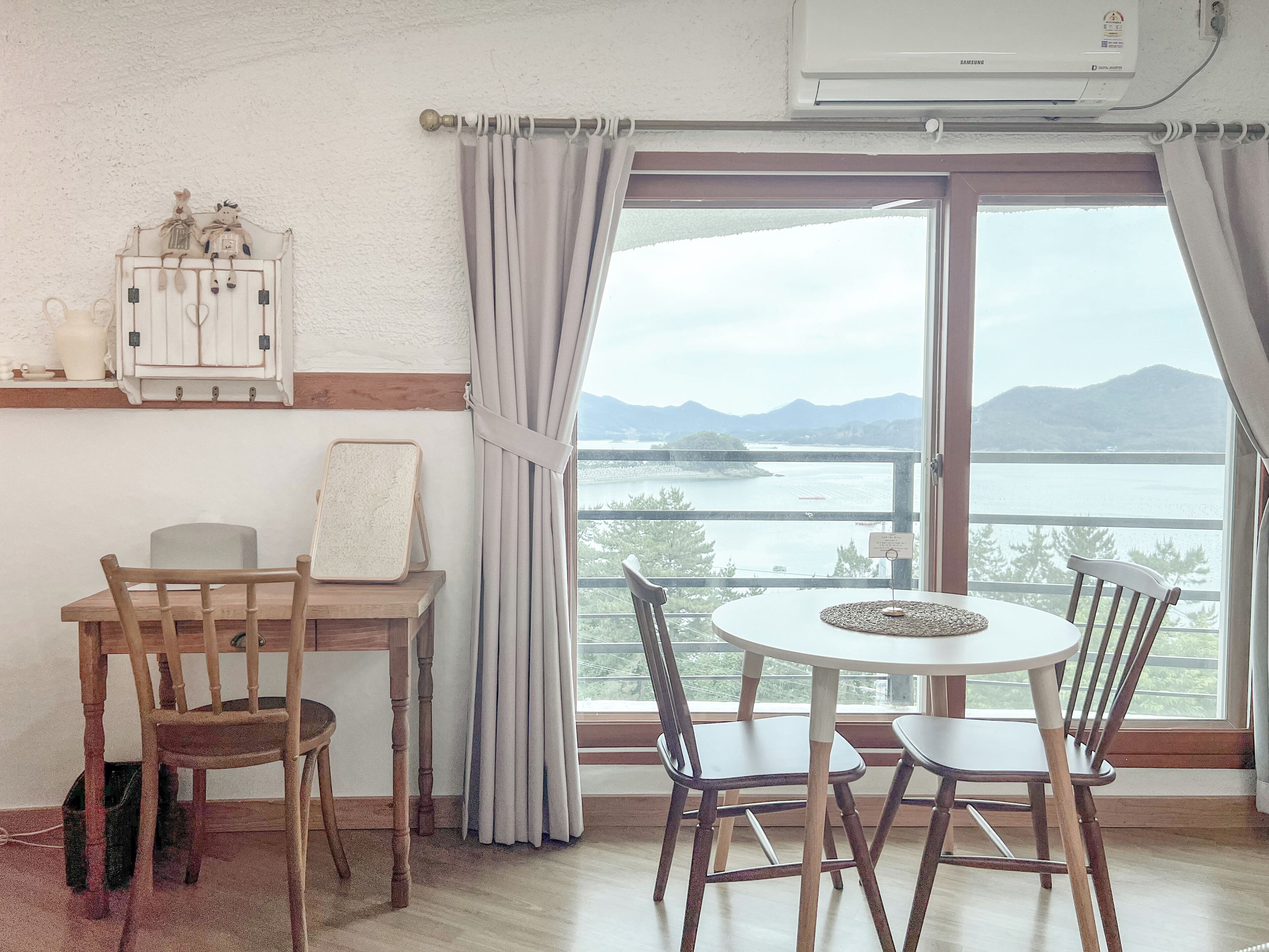 A room with a wooden dining table and chairs, a white air conditioning unit, a view of a body of water and mountains through a large window with curtains.